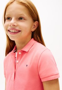 Χαμογελαστή νεαρή κοπέλα με μακριά κόκκινα μαλλιά, που φοράει ροζ πουκάμισο με γιακά, γυρισμένη ελαφρώς προς τα αριστερά σε ένα απλό λευκό φόντο.