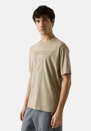 Joven con camiseta beige de manga corta con un logo triangular en relieve y pantalones grises, de pie frente a un fondo liso.