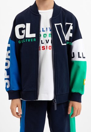 Niño con chaqueta azul marino con cremallera y bloques de color azul, verde y blanco y texto, encima de una camiseta blanca con letras multicolores.