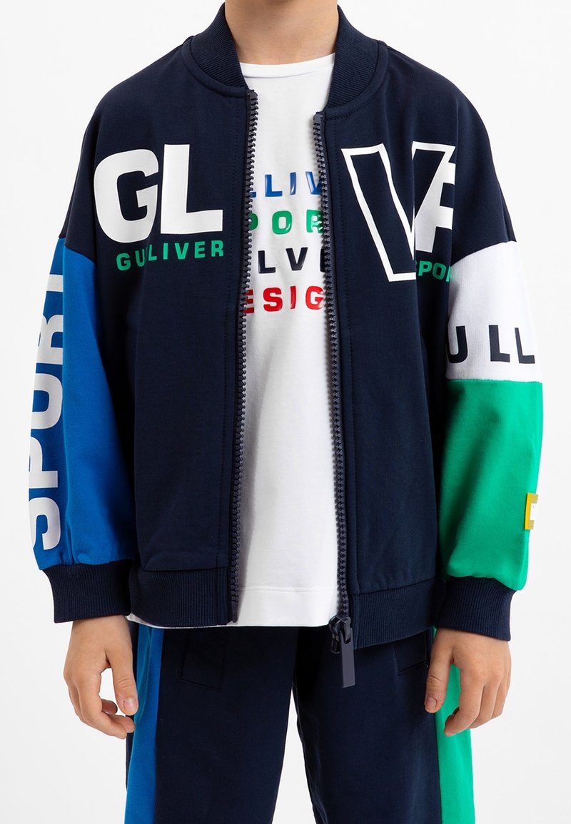 Barn iført en marineblå lynlåsjakke med blokfarver i blå, grøn og hvid samt tekst, over en hvid skjorte med flerfarvede bogstaver.