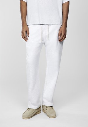 Moitié inférieure d'une personne portant un pantalon blanc à cordon, des chaussures en daim beige et une chemise à manches courtes gris clair sur un fond uni.