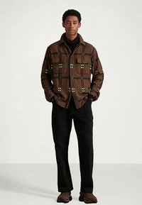 Moški oblečen v rjavo karo srajco na gumbe, pod katero nosi črn polo, s črnimi hlačami in rjavimi čevlji, stoji pred enobarvno svetlo ozadje.