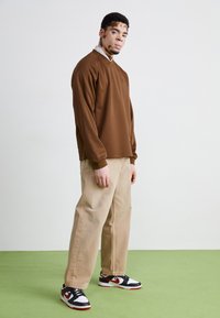 Haut marron à manches longues avec décolleté en V, pantalon beige à larges jambes et baskets noir/rouge avec accents blancs. Avec un fond clair.