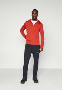 Camisola com fecho de correr vermelha, com gola alta, combinada com calças pretas. O conjunto apresenta um ajuste justo e um tecido texturizado. Ténis de caminhada completam o visual.