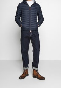 Giacca puffy navy con cappuccio, chiusura con zip e accento arancione. Abbinata a jeans scuri e stivaletti marroni con lacci e orli risvoltati.