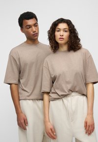 Δύο άτομα φορούν ταίριαστα, φαρδιά μπλουζάκια με στρογγυλή λαιμόκοψη σε ανοιχτό καφέ χρώμα, συνδυασμένα με άνετα, ανοιχτό μπεζ παντελόνια.