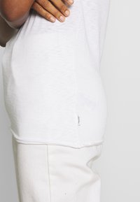 Camiseta blanca de manga corta que muestra un ajuste relajado y una textura de tela suave. Presenta una pequeña etiqueta en la costura lateral, combinada con pantalones blanco roto.