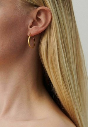 Pikajuukseline naine, kes kannab vasakul kõrval keskmise suurusega kuldset rõngakõrvarõngast, neutraalse taustaga.