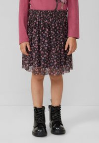 Kind, das einen schwarzen, blumigen Rock, ein pinkes langärmliges Oberteil und glänzende schwarze Schnürstiefel trägt, steht vor einem schlichten Hintergrund.