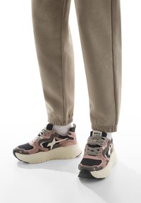 Sneakers con un design rosa e nero, caratterizzati da un accento a stella bianca, tomaia testurizzata e suola bianca spessa. Abbinati a pantaloni jogger beige.