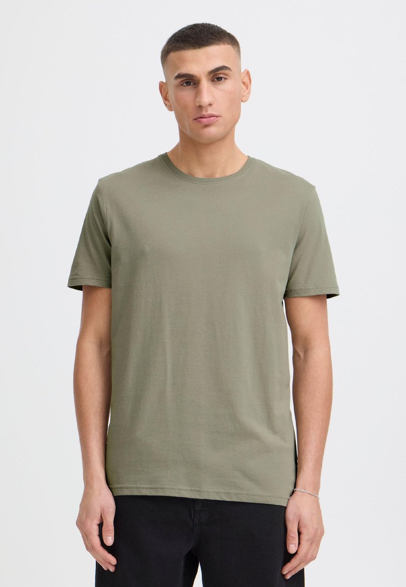 Junger Mann mit kurzen Haaren, der ein olivgrünes kurzärmeliges T-Shirt und schwarze Hosen vor einem schlichten hellen Hintergrund trägt.