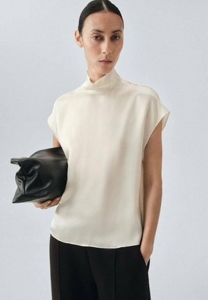 Blusa in seta crema, collo alto, maniche corte. Abbinata a una pochette in pelle nera. Tessuto liscio, vestibilità comoda. Visibili pantaloni scuri.