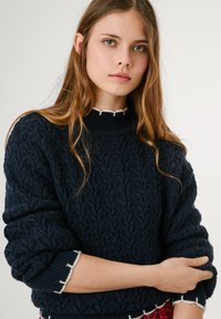 Jūras spēku kabeļu adījuma džemperis ar augstu ribotu apkakli un teksturētu rokdarbību. Izceļas ar baltām viļņainām akcentiem apkaklē un piedurknēs.