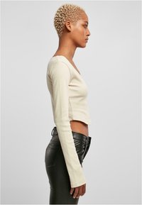 Hosszú ujjú, világos bézs bordázott anyagú cropped top; V-nyakú kivágással rendelkezik. Feszes, fekete bőrnadrággal párosítva.