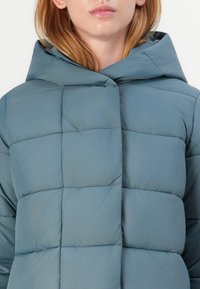 Pěstovaný kabát v tlumené modro-zelené barvě s velkou kapucí, vybavený prošívaným designem a viditelným šitím. Měkká textura, minimalistický styl.