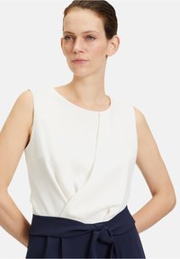 Fehér ujjatlan blúz, rakott részletekkel és kerek nyakkivágással, mélykék szoknyával párosítva, amelynek derekán megkötős öv található.