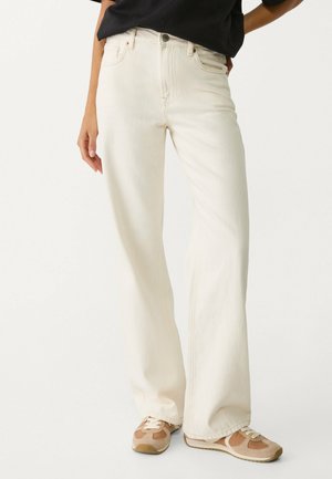 Kvinde iført højtaljede, vidde, off-white jeans og beige sneakers, stående med en hånd hvilende på hoften.