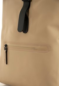 Beige vanntett ryggsekk med glatt tekstur, med svart spenne, gjennomsiktig glidelåslomme og preget logo på overflaten.