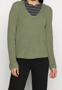 Lichtgroene gebreide trui met een V-hals en lange mouwen, gedragen over een zwart-wit gestreept shirt. Gevlochten stof en een ontspannen pasvorm.