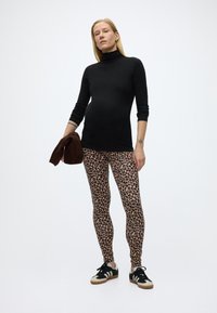 Γυναίκα που στέκεται φορώντας κολάν με λεοπάρ πατρόν, μαύρο ζιβάγκο και αθλητικά παπούτσια, κρατώντας μια διπλωμένη καφέ ζακέτα μπροστά σε απλό λευκό φόντο.