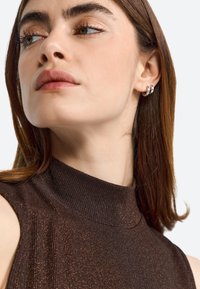 Brown sleeveless top with a high neck, textured ribbed fabric, and metallic shimmer accents. Model adorned with small silver hoop earrings.