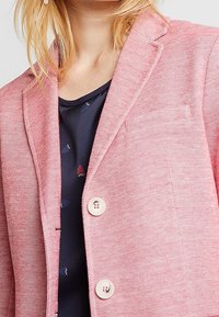 Blazer in misto lino rosa chiaro con superficie testurizzata, rever a lancia e due bottoni bianchi. Presenta una tasca sul petto.