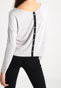 Szary sweter o fakturowanej powierzchni z szerokim dekoltem i czarnymi guzikami z tyłu. Długie rękawy i krótki fason. Noszone są czarne spodnie.