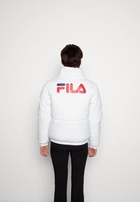 Hvid dunjakke med høj krave, med en rød og blå FILA-logo på ryggen, kontrasteret mod en ensfarvet baggrund.
