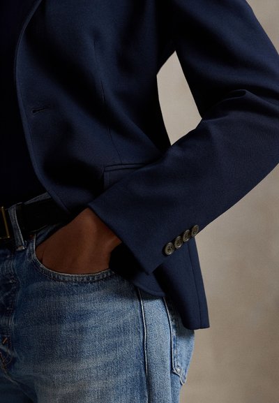 Άτομο που φοράει navy σακάκι και μπλε τζιν με το ένα χέρι χαλαρά στην τσέπη σε ουδέτερο φόντο.