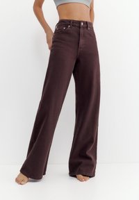 Person iført højtaljede, vidde mørkebrune jeans og en lysegrå crop top, stående barfodet mod en ensfarvet hvid baggrund.