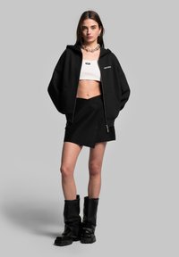 Felpa nera oversize con zip, top bianco corto, gonna avvolgente nera e stivali neri al ginocchio. Caratterizzata da una collana con catena argentata.