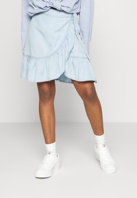 Saia envolvente em denim azul claro com uma bainha de volantes em camadas, com detalhe de laço lateral, combinada com ténis brancos e meias até ao tornozelo.