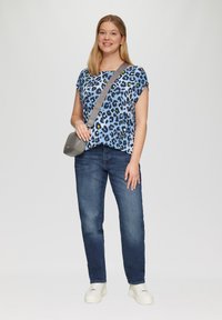 Hellblaues Leopardenmuster-T-Shirt mit kurzen Ärmeln, kombiniert mit dunkelblauen Jeans und weißen Sneakers. Inklusive einer grauen Umhängetasche.