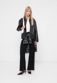 Veste en cuir noir de style aviateur, chemise blanche, chemise à carreaux nouée autour de la taille, pantalons larges noirs et escarpins noirs. Détail du sac à main blanc.