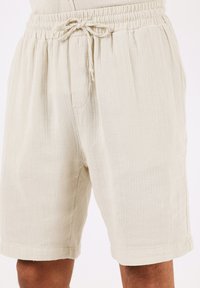 Pantaloni corti beige chiaro realizzati in tessuto testurizzato con vita elastica e cordino. Presenta tasche laterali e un design dalla vestibilità rilassata.