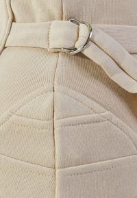 Tissu beige texturé avec des coutures diagonales, une silhouette cintrée et un détail de boucle en argent sur une large ceinture.