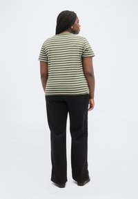 Γυναίκα με πλεξούδες που φοράει ριγέ μπλουζάκι σε ελιά πράσινο και άσπρο και μαύρο παντελόνι, στέκεται με την πλάτη στραμμένη σε απλό φόντο.