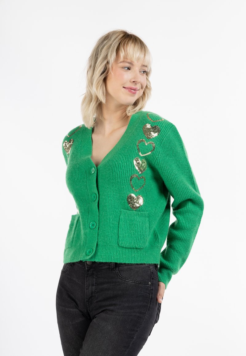 Cardigan de punto verde con cuello en V, cinco botones, dos bolsillos frontales y lentejuelas doradas en forma de corazón como acentos. Textura suave con detalles acanalados.