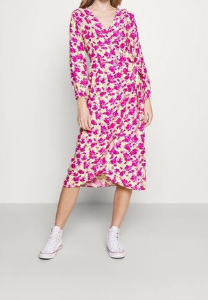 Frau trägt ein gelbes Wickelkleid mit leuchtend pinkem Blumenmuster und langen Ärmeln, kombiniert mit weißen High-Top-Sneakers.