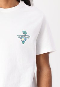T-shirt en coton blanc avec un logo triangulaire multicolore "GUESS JEANS" comprenant un palmier et un design de soleil sur la poitrine gauche. Manches courtes.