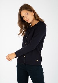 Granatowy kardigan wykonany z miękkiego materiału, z okrągłym dekoltem, długimi rękawami, złotymi guzikami i małą naszywką z logo u dołu.