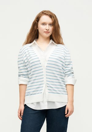 Vrouw met lang golvend haar, een witte blouse en een witte cardigan met lichtblauwe strepen, gecombineerd met donkerblauwe spijkerbroek, staand met het gezicht naar voren.