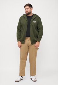 Maslinasto zelena zip-up dukserica s konopcem, crna majica ispod, u kombinaciji s bež hlačama i bijelim teniscima s crnim prugama.