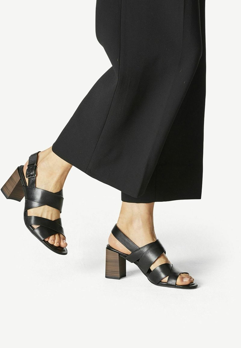 Personne portant un pantalon noir à jambes larges et des sandales en cuir noir à talons blocs avec brides à la cheville, soulevant légèrement un pied du sol.