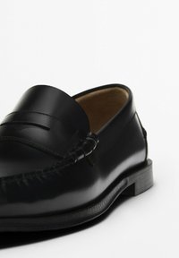 Black leather loafers with a smooth finish, featuring a decorative strap and stitching detail, and a textured sole for grip.