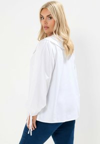 Biała bluzka z dużym kołnierzem, z puffowymi długimi rękawami i wiązaniami przy mankietach. Gładka faktura, luźny krój. Połączona z niebieskimi dżinsami.