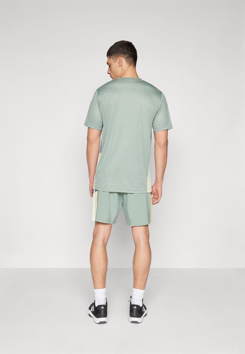 Camiseta deportiva verde claro y pantalones cortos con acentos en amarillo, de manga corta y corte relajado. Combinados con zapatillas deportivas negras y calcetines blancos.