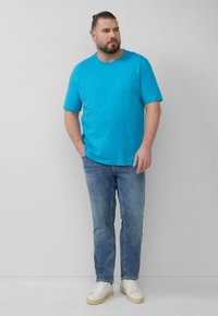 Turquoise T-shirt met korte mouwen en een borstzak, gecombineerd met lichtblauwe jeans en witte sneakers. Simpel, casual outfitontwerp.