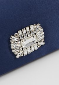 Broche rectangulaire en argent avec des cristaux clairs disposés sur un fond en tissu bleu foncé.