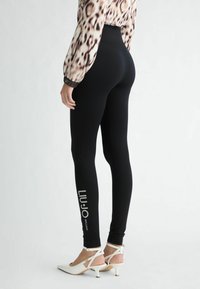 Leggings negros de cintura alta, con un estampado de logo en el lado izquierdo; combinados con zapatos de tacón alto, abiertos por delante y de color blanco.
